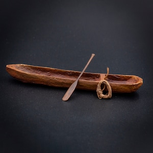 Puede incluir: Un canoa de madera en miniatura con un pequeño remo de madera y una cuerda marrón atada en un círculo. El canoa y el remo son marrones y la cuerda es marrón claro.