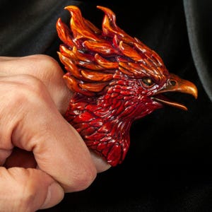 Op de afbeelding: Een gedetailleerde, handgeschilderde fenikssculptuur in tinten rood, oranje en goud. De kop van de vogel is het middelpunt, met ingewikkelde veerdetaillering en een open snavel. De sculptuur wordt in een hand gehouden tegen een donkere achtergrond.