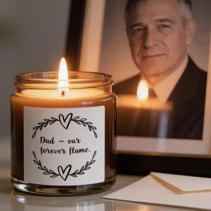 May include: A lit candle in a glass jar with a white label that reads "Dad - our forever flame." The candle sits in front of a framed black and white portrait and a white envelope.