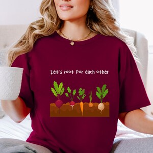 Può includere: Una t-shirt a maniche corte color bordeaux con una grafica di vari ortaggi a radice che crescono in terra marrone. Sopra le verdure, il testo bianco recita, Let's root for each other. La maglietta ha un girocollo e una vestibilità casual.