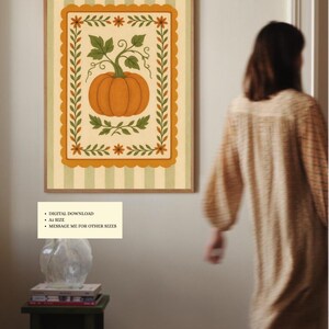 Puede incluir: Una impresión artística enmarcada con una vibrante calabaza naranja con hojas y vides verdes, sobre un fondo crema con un borde decorativo. La obra de arte está enmarcada en un marco de madera y tiene un fondo a rayas.
