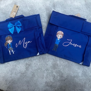 May include: Two royal blue book bags with top handles and flaps. One bag features a blue satin bow, a cartoon illustration of a girl, and the name Mya in white script. The other bag features a cartoon illustration of a boy and the name Jenson in white script.