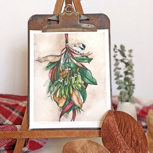 watercolor art print of magnolia leaves and mistletoe hanging from a burgundy ribbon with chickadee bird perched on top. art print is clipped to a vintage clipboard and standing on a table-top easel with plaid blanket and magnolia leaves at base