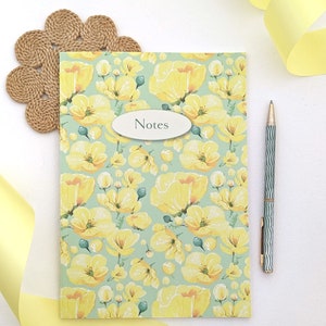 Puede incluir: Una libreta verde con un patrón floral amarillo y la palabra "Notes" en la portada. La libreta está sobre una superficie blanca con una cinta amarilla y un posavasos tejido marrón.