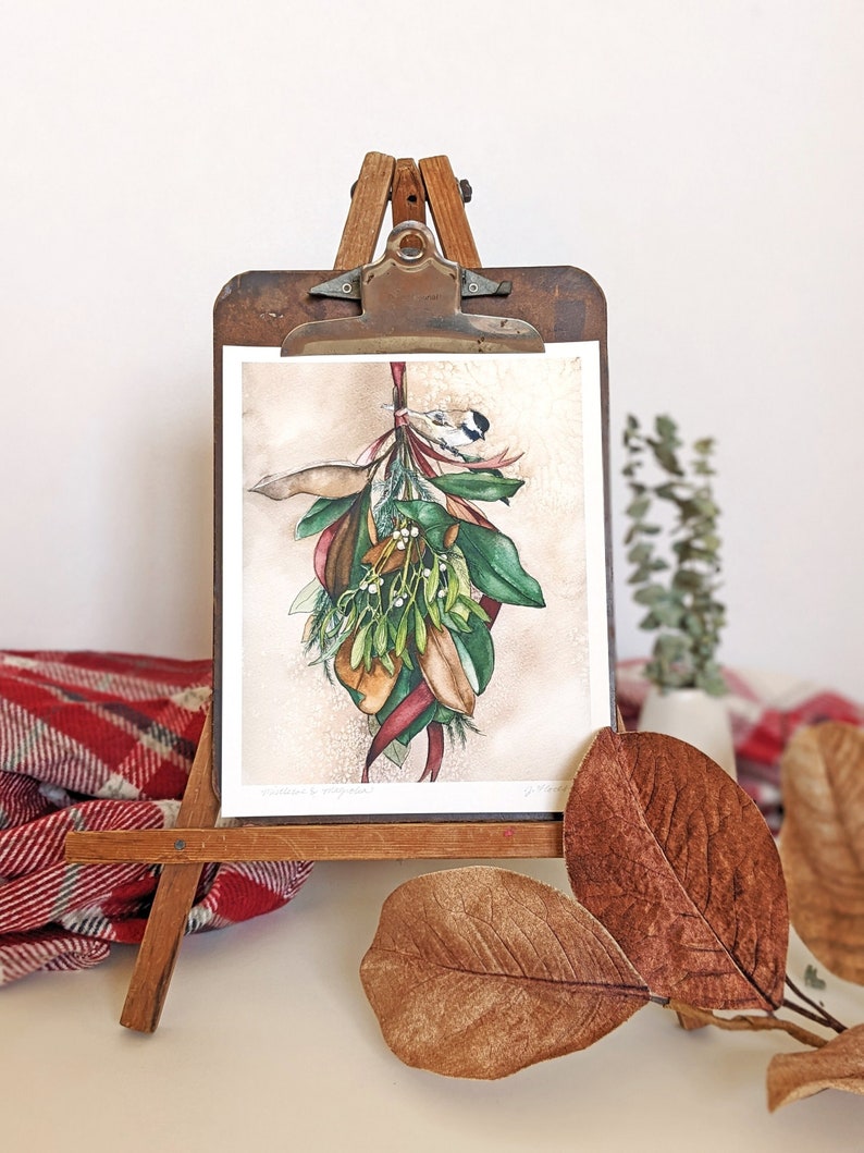 watercolor art print of magnolia leaves and mistletoe hanging from a burgundy ribbon with chickadee bird perched on top. art print is clipped to a vintage clipboard and standing on a table-top easel with plaid blanket and magnolia leaves at base