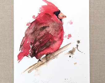 May include: Watercolor painting of a red cardinal bird perched on a branch. The bird is painted in shades of red, brown, and black. The background is white with a few brown splatters of paint.