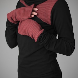 Puede incluir: Una camisa negra de manga larga con un panel rojo de canalé a través del pecho y los hombros. La camisa tiene un cuello alto y una textura de canalé. La modelo lleva un par de guantes sin dedos de canalé rojos.