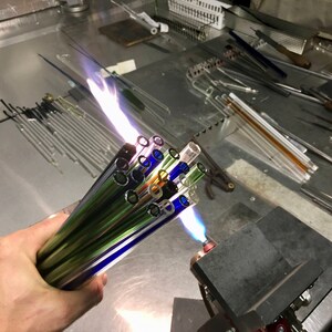 A hand is holding a bundle of colorful glass rods, which are being heated by a flame. The rods are arranged in a bundle, and the flame is burning brightly, creating a vibrant display of colors.