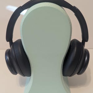 May include: Black over-ear headphones resting on a light green head-shaped display stand. The headphones have large earcups and a padded headband. The stand is a solid color and has a smooth surface.