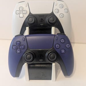 Puede incluir: Dos mandos de juego inalámbricos, uno blanco y otro morado oscuro, apilados en un soporte de carga blanco y negro. Los mandos tienen botones negros y almohadillas direccionales. El soporte tiene el logotipo de PlayStation.