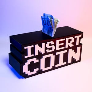 以下が含まれることがあります： 白のピクセル化された文字で「INSERT COIN」と書かれた黒い長方形の貯金箱。箱の中にはお札が見えます。貯金箱はレトロなビデオゲームの美学を持っています。