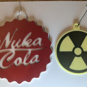 Può includere: Due ornamenti decorativi. Uno è un cerchio rosso con bordo smerlato con il testo bianco "Nuka Cola". L'altro è un cerchio giallo con un simbolo di radiazione nero. Entrambi hanno anelli per appenderli.