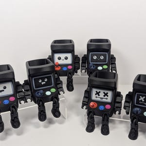 Puede incluir: Cinco macetas negras con forma de robot con diferentes expresiones faciales y botones de colores. Las macetas están dispuestas en fila sobre una superficie blanca.