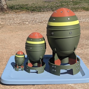May include: Three olive green, red, and yellow decorative bomb models of varying sizes are displayed on a light blue tray. The models have a rounded shape with a cylindrical base and are designed with rivets and a circular base.