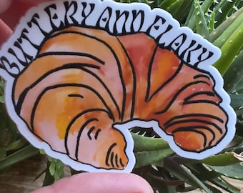 May include: A sticker depicting a watercolour illustration of a croissant in orange and brown hues, with the words "BUTTERY and FLAKY" in a black, stylized font. The sticker is held in front of a backdrop of green plants.