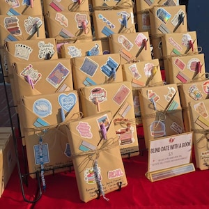 May include: A selection of 'blind date with a book' packages, wrapped in brown paper and decorated with stickers and twine. The display is on a tiered black metal stand, with a red cloth covering the table.