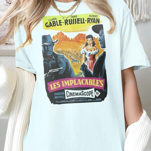 May include: Light blue t-shirt with a vintage movie poster print featuring Clark Gable, Jane Russell, and Robert Ryan in "The Unforgiven". The poster is in color and features the title "Les Implacables" and "Cinemascope".