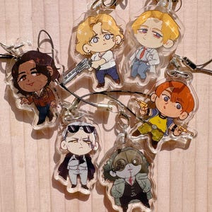 May include: A collection of six clear acrylic charms featuring chibi-style illustrated characters. Each charm includes a black cord loop and metal clasp. The characters include figures with various hair colors, outfits, and accessories like guns and masks.