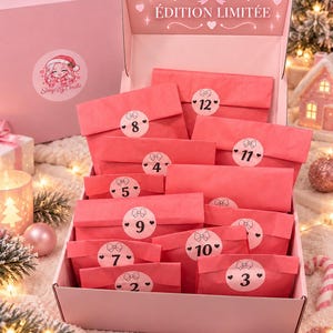 Puede incluir: Una caja de cartón rosa para calendario de adviento que contiene doce sobres de papel rojo, cada uno sellado con una pegatina circular blanca con un número negro del 2 al 12 y un icono de lazo. La tapa muestra el texto ÉDITION LIMITÉE con un lazo.
