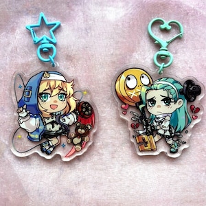 Könnte beinhalten: Zwei Acryl-Schlüsselanhänger mit Charakteren im Anime-Stil. Einer zeigt einen blonden Charakter in einem blauen Hoodie, der einen Teddybären und ein Schwert hält. Der andere zeigt einen Charakter mit türkisfarbenem Haar und einem Schlüssel.