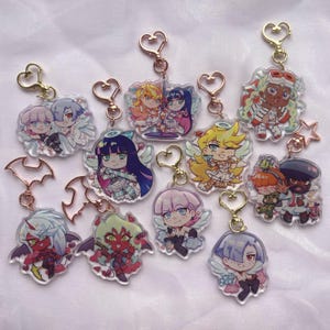 Könnte beinhalten: Eine Sammlung von zehn durchsichtigen Acryl-Schlüsselanhängern mit farbenfrohen Illustrationen im Anime-Stil. Jeder Anhänger ist an einem goldfarbenen Metallverschluss in verschiedenen Formen wie Herzen und Sternen befestigt und zeigt diverse Figuren mit Flügeln, Hörnern und lebhaften Outfits.