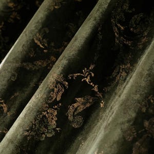 Puede incluir: Primer plano de tela verde oscuro con patrones florales dorados. La tela tiene un acabado envejecido y texturizado, con pliegues que crean sombras. Los patrones dorados son un diseño floral y de volutas repetido.