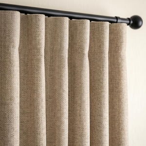 Puede incluir: Cortinas de color beige con un sutil patrón de espiga que cuelgan de una barra de metal negro mate con remates redondos. La tela tiene una apariencia texturizada con pliegues verticales que crean ondas uniformes en todo el cortinaje.