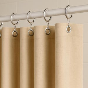 Puede incluir: Cortinas de tela beige colgadas de una barra blanca con anillos metálicos plateados. Las cortinas están hechas de un material texturizado y están parcialmente cerradas, creando pliegues suaves. La paleta de colores neutros sugiere un diseño simple y elegante.