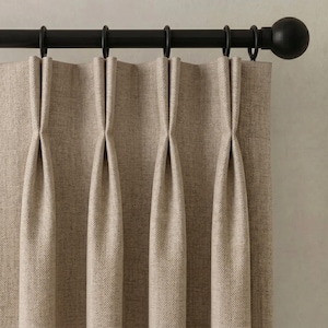Op de afbeelding: Een beige gordijn met een subtiel visgraatpatroon en diepe plooien. De stof hangt aan een matzwarte metalen gordijnroede met bijpassende ronde ringen en een grote bolvormige knop aan het uiteinde van de roedeconstructie.
