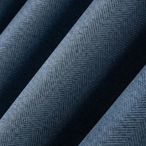 May include: Close-up of a textured, blue fabric with a herringbone pattern. The fabric is folded, creating diagonal lines and shadows. The weave is visible, showing the detailed texture of the material. The colour is a deep, muted blue.