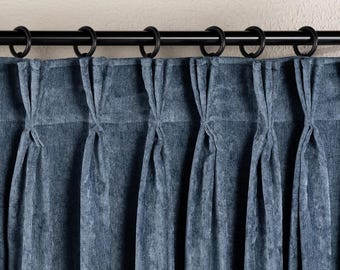 May include: A blue velvet curtain featuring a pinch pleat header style, suspended from a black metal curtain rod by matching black circular rings. The fabric has a soft, textured appearance with vertical folds creating deep, uniform pleats.