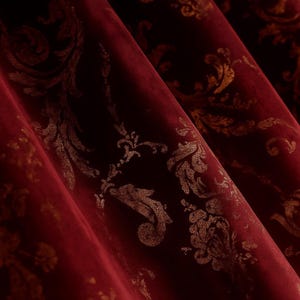 May include: Close-up of a deep red velvet fabric with a gold baroque pattern. The fabric is draped in soft folds, revealing the texture and sheen of the material. The ornate design features swirling floral and leaf motifs.