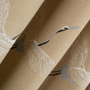 Pode incluir: Close-up de tecido bege com garças brancas bordadas em voo. As garças têm detalhes em preto e azul claro. O tecido está dobrado, mostrando a textura e os detalhes do bordado.