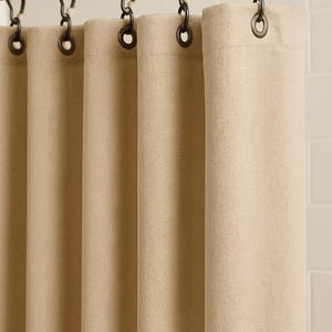 Puede incluir: Cortina de ducha beige con tejido texturizado, colgada de una barra de metal oscuro con ganchos a juego. La cortina est&aacute; hecha de un material grueso y opaco, que proporciona privacidad. El fondo es una pared de azulejos blancos.