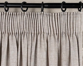 May include: Close-up of a beige curtain panel with pleats hanging from a black curtain rod with black rings. The curtain has a textured appearance and hangs in vertical folds.