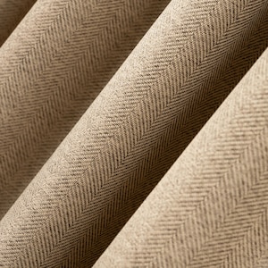 Peut inclure: Gros plan sur un tissu beige avec un motif en chevrons. Le tissu est plié, créant des lignes diagonales et des ombres. Le tissage est visible, montrant une surface texturée. La couleur est un beige neutre avec des mouchetures plus foncées.