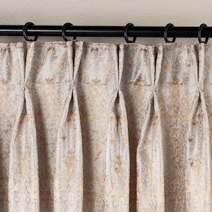 May include: Close-up of a beige curtain with a gold floral pattern. The curtain is hanging from a black rod with black rings. The fabric is pleated, creating vertical folds.