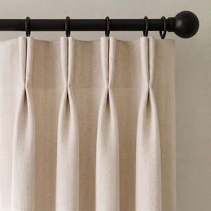 Puede incluir: Panel de cortina de color blanco roto con un sutil patrón de tejido en espiga. La tela está suspendida de una barra de cortina de metal negro mate mediante cuatro anillas circulares, creando pliegues uniformes que caen verticalmente.