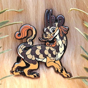 Puede incluir: Un pin de esmalte dorado, marrón y naranja de una criatura mítica con cola enroscada, cuernos y escamas. La criatura está representada en un estilo de dibujos animados con una expresión amigable.