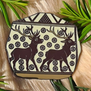 Pode incluir: Um broche de esmalte branco com um design preto e castanho de dois veados em pé sobre um fundo estampado. O fundo é de cor amarelo claro com círculos pretos e um padrão geométrico.