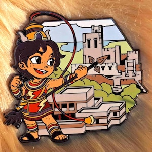 Peut inclure: Un personnage de dessin animé aux cheveux bruns et vêtu d'une tenue rouge et jaune tient une lance et un lasso. Le personnage se tient devant un bâtiment avec un château en arrière-plan.