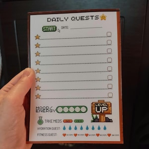 Może przedstawiać: Prostokątny notatnik z pikselowym wzorem, zatytułowany "DAILY QUESTS". Zawiera sekcje do śledzenia codziennych zadań, nastroju i nawodnienia. Notatnik ma drewnianą ramkę i zawiera tekst "START", "TAKE MEDS" i "LEVEL UP".
