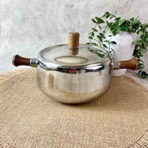 May include: A stainless steel cooking pot with a polished finish, featuring two dark brown wooden side handles and a matching wooden knob on the lid. The pot has a rounded body and a flat lid, designed for stovetop use in a kitchen setting.