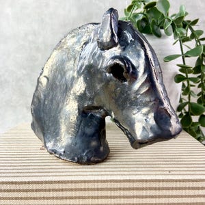 May include: A ceramic horse head sculpture with a glossy, dark metallic finish featuring iridescent blue and gold highlights. The piece has a textured, hand-moulded surface with a defined ear, eye socket, and snout, capturing the profile of a horse.