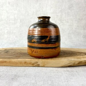 May include: A small, round ceramic vase featuring a glossy finish with horizontal bands of dark brown and earthy orange. The vessel has a narrow neck and a wide, rounded body, with a small, circular maker's mark stamped near the base.