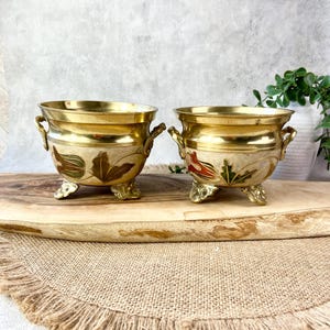 May include: Two matching brass planters with a polished gold-tone rim and handles. The lower section features an off-white enamel finish decorated with raised, hand-painted floral motifs in shades of green, red, and brown, supported by three feet.