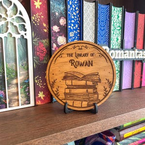 Op de afbeelding: Een rond houten bord met de tekst "THE LIBRARY OF: ROWAN" en een illustratie van opgestapelde boeken. Het bord staat op een zwarte standaard, met een boekenplank en boeken op de achtergrond.
