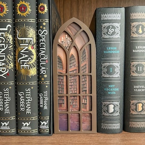 May include: A decorative bookshelf window with a library scene, placed among several hardback books. The books have dark covers with gold lettering and accents. The window is a light brown color.