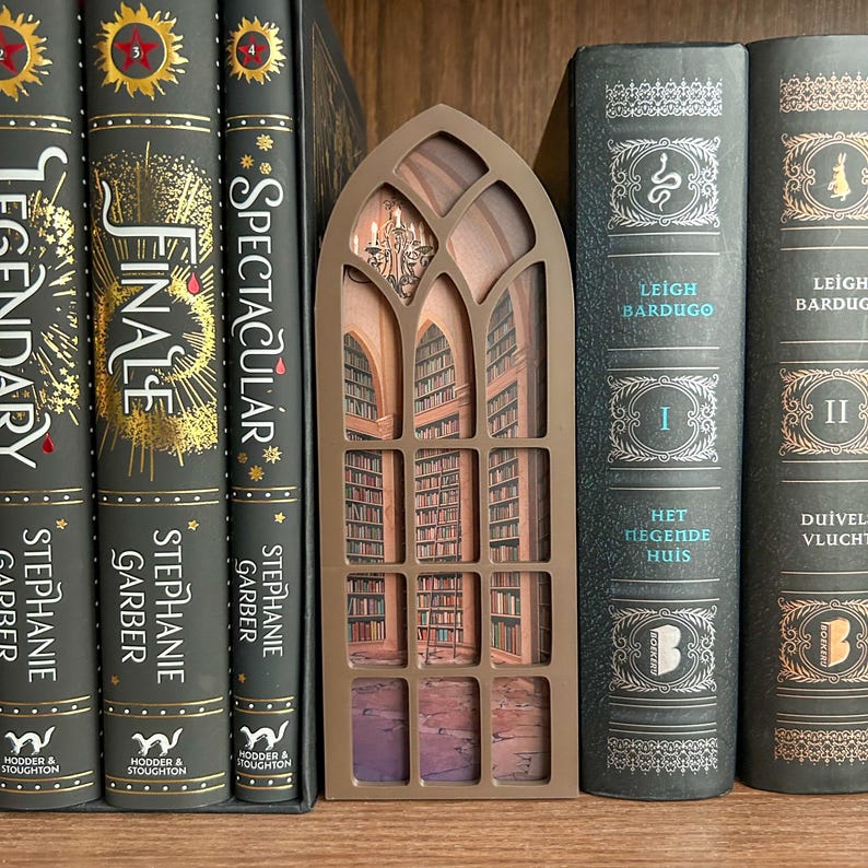 May include: A brown, arched book nook insert featuring a detailed, multi-layered illustration of a library interior with tall bookshelves, a rolling ladder, and a chandelier, positioned between several hardcover books on a wooden shelf.
