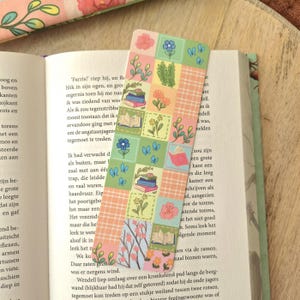Op de afbeelding: Een rechthoekige boekenlegger met een rasterpatroon van pastelkleurige vierkanten. Het ontwerp bevat illustraties van stapels boeken, open boeken, diverse bloemen, groen gebladerte en kleine blauwe vlinders tegen een lichtgroene en ruitjesachtergrond.